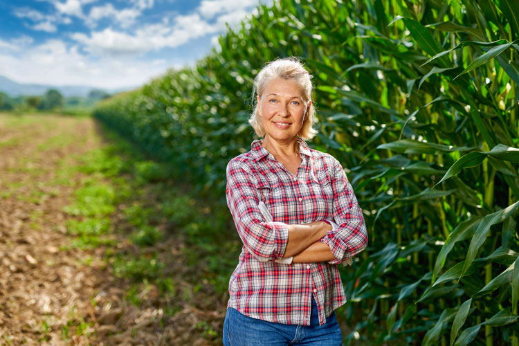 Older woman standing on the edge of a corn field with her arms crossed.